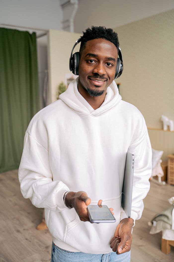 A cheerful man wearing headphones, holding a smartphone and laptop indoors.